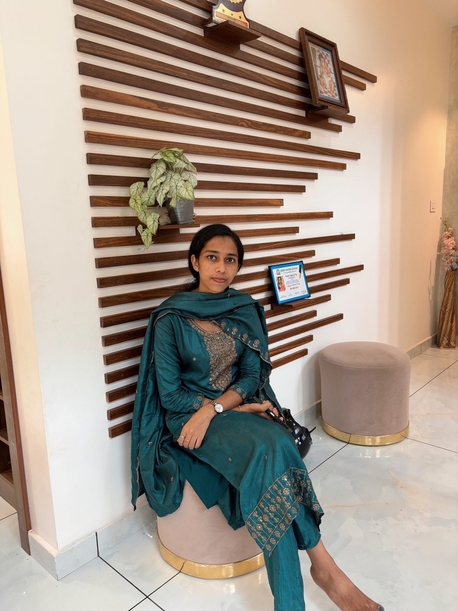 Woman in a teal traditional outfit sitting indoors against a decorative wooden wall.
